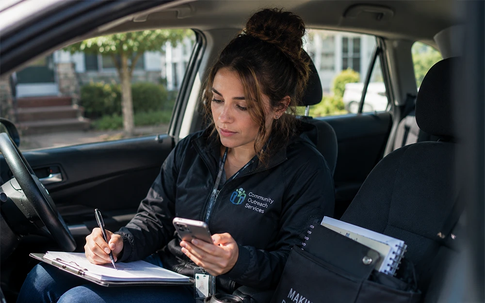 Community outreach worker sitting in a parked vehicle completing paperwork and checking a smartphone before a field visit, representing lone worker safety and mobile social assistance services.